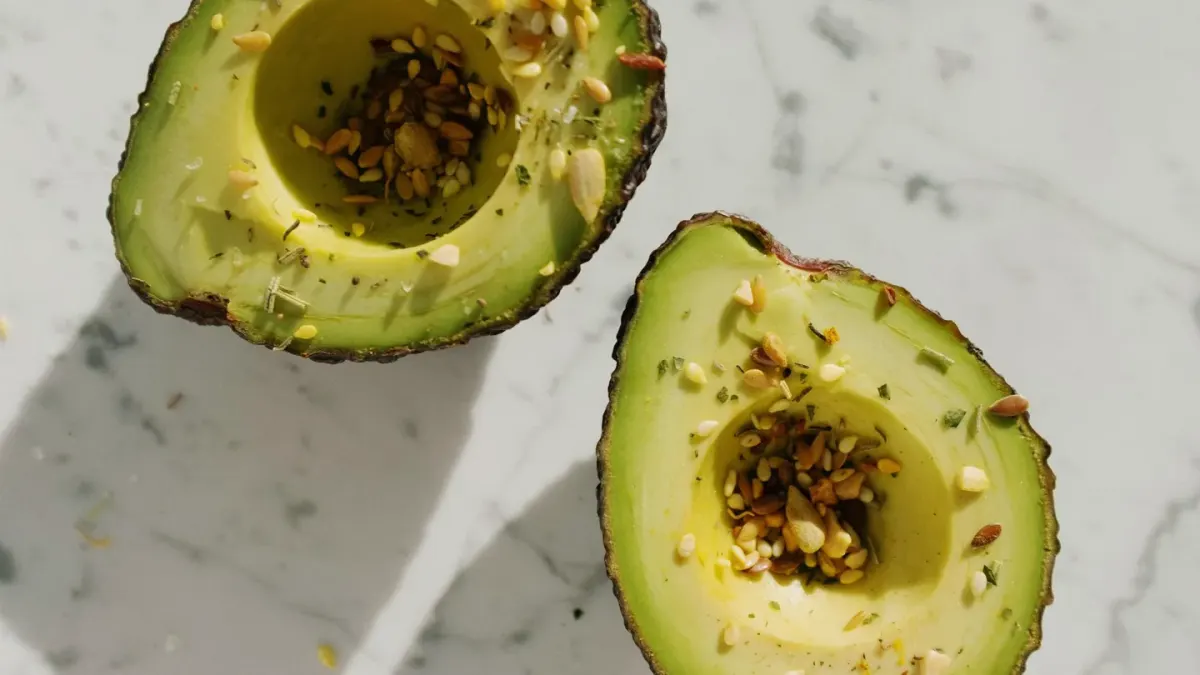 A halved avocado with the pit exposed sitting on a kitchen counter next to a dog sniffing nearby